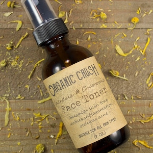 May include: A brown glass bottle of Organic Crush Face Toner with a black spray top. The label reads "Calendula & Chamomile Face Toner" and lists benefits. The bottle is 2 oz. and is suitable for all skin types. The background is a wooden surface with dried flowers.