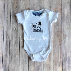May include: White baby onesie with black embroidered text that reads "babe lincoln" with a small black top hat above the word "babe". The onesie has a white tag that reads "Stitched".