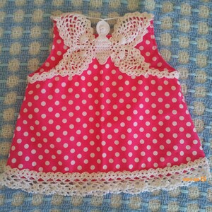 Könnte beinhalten: Ein pinkfarbenes, ärmelloses Kleid mit weißen Punkten und einem weißen gehäkelten Schmetterlingsmuster am Ausschnitt. Das Kleid hat einen weißen gehäkelten Besatz am Ausschnitt und Saum.