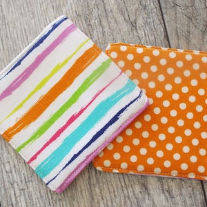 May include: Two square fabric coasters. One coaster features a white background with diagonal multi-colored brushstroke stripes in pink, orange, yellow, teal, and dark blue. The second coaster is orange with a repeating pattern of small white polka dots.