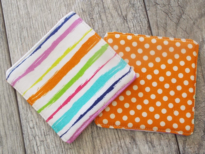 May include: Two square fabric coasters. One coaster features a white background with diagonal multi-colored brushstroke stripes in pink, orange, yellow, teal, and dark blue. The second coaster is orange with a repeating pattern of small white polka dots.