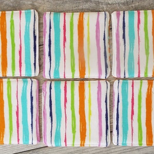 May include: A set of six square fabric coasters featuring a white background with vertical, brushstroke-style stripes in vibrant shades of orange, lime green, turquoise, magenta, and dark blue. Each coaster has stitched edges and a soft, flat surface.
