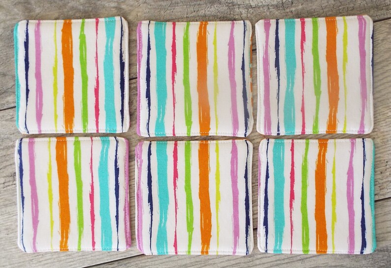 May include: A set of six square fabric coasters featuring a white background with vertical, brushstroke-style stripes in vibrant shades of orange, lime green, turquoise, magenta, and dark blue. Each coaster has stitched edges and a soft, flat surface.