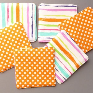 May include: A set of five square fabric coasters featuring two distinct patterns. Three coasters display a bright orange background with white polka dots, while two coasters feature a white background with vertical or horizontal multicolored brushstroke stripes.