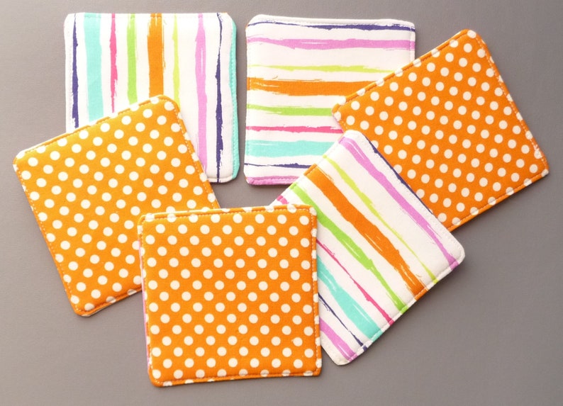 May include: A set of five square fabric coasters featuring two distinct patterns. Three coasters display a bright orange background with white polka dots, while two coasters feature a white background with vertical or horizontal multicolored brushstroke stripes.