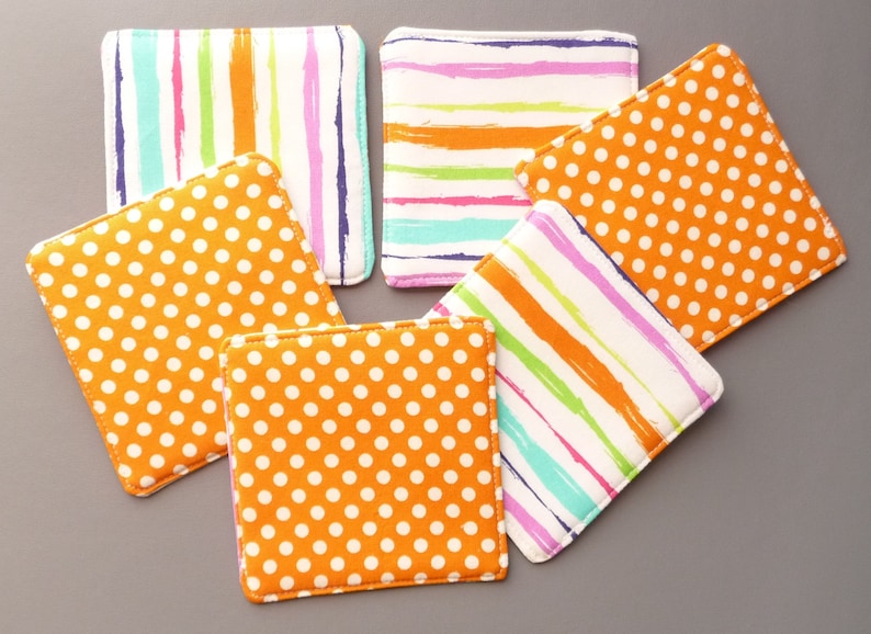 May include: A set of five square fabric coasters featuring two distinct patterns. Three coasters display a white background with orange polka dots, while two coasters feature a white background with multicolored vertical or horizontal stripes.