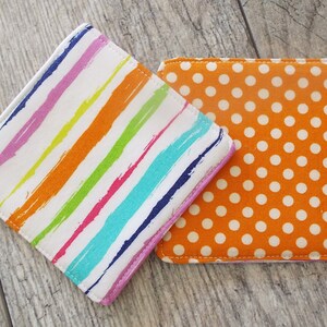 May include: Two square fabric coasters. One coaster features a white background with diagonal, multi-colored brushstroke stripes in purple, orange, lime green, and teal. The second coaster is orange with a repeating pattern of small white polka dots.