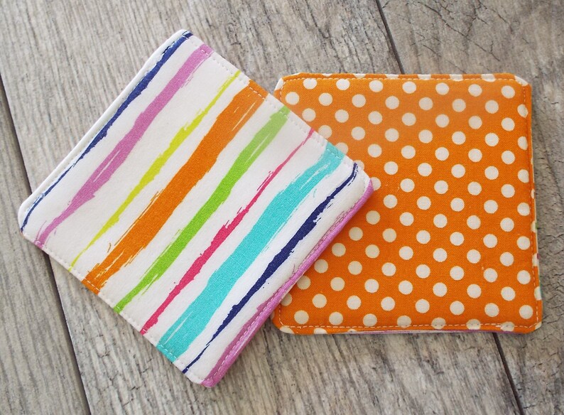 May include: Two square fabric coasters. One coaster features a white background with diagonal, multi-colored brushstroke stripes in purple, orange, lime green, and teal. The second coaster is orange with a repeating pattern of small white polka dots.