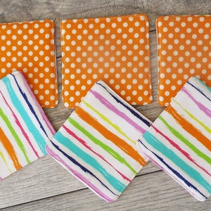 May include: A set of six square fabric coasters. Three coasters feature an orange background with a white polka dot pattern. The other three coasters feature a white background with diagonal stripes in orange, pink, green, and blue colors.