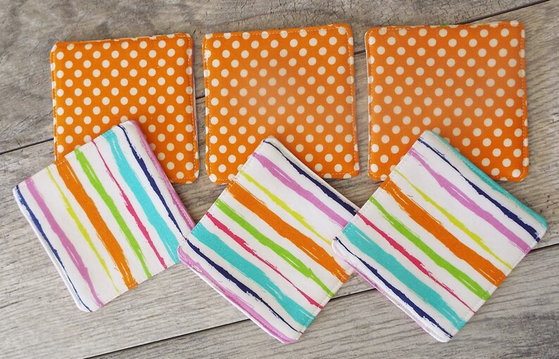 May include: A set of six square fabric coasters. Three coasters feature an orange background with a white polka dot pattern. The other three coasters feature a white background with diagonal stripes in orange, pink, green, and blue colors.