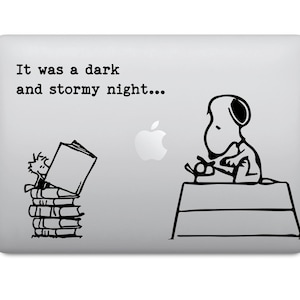 Puede incluir: Un ordenador portátil con un diseño de dibujos animados en blanco y negro. El diseño presenta a Snoopy sentado en una caseta para perros, un personaje leyendo un libro sobre una pila de libros y el texto "It was a dark and stormy night..."