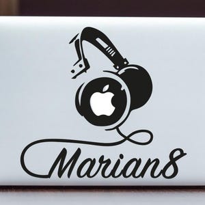 Könnte beinhalten: Ein schwarz-weißer Aufkleber mit Kopfhörern und einem Apple-Logo auf einem weißen Laptop. Der Text "Marians" befindet sich unter den Kopfhörern.