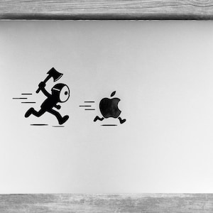 Può includere: Un coperchio per computer portatile color argento con un adesivo in vinile nero che raffigura una figura mascherata con un'ascia che corre verso un logo Apple nero che sembra scappare, con linee di movimento orizzontali per entrambi.