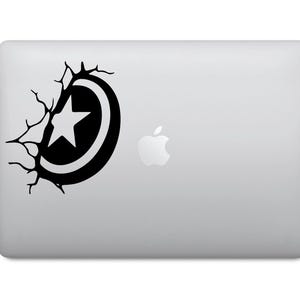 Peut inclure: Autocollant noir et blanc représentant un bouclier avec une étoile, donnant l'impression de percer une surface. L'autocollant est appliqué sur un ordinateur portable argenté, avec le logo Apple visible.