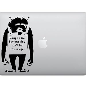 Könnte beinhalten: Ein schwarz-weißer Aufkleber mit einem Affen, der ein Schild trägt, auf dem steht "Laugh now but one day we'll be in charge". Der Aufkleber befindet sich auf einem silbernen Laptop.