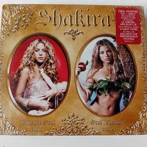 May include: A gold-coloured box featuring a picture of Shakira on the front. The box contains two DVDs, "Fijación Oral, Vol. 1" and "Oral Fixation, Vol. 2".