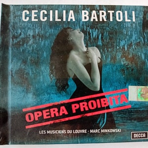 Puede incluir: Una portada de CD con una mujer de cabello oscuro y un vestido negro, cantando sobre un fondo azul y verde. El título "Opera Proibita" está escrito en letras rojas y blancas. El álbum es de Cecilia Bartoli, Les Musiciens du Louvre y Marc Minkowski.