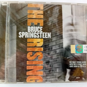 Puede incluir: Una caja de CD con un fondo negro y naranja que presenta el texto "THE RISING" en letras grandes, en negrita y de color naranja. El texto "BRUCE SPRINGSTEEN" está en letras blancas sobre el título principal. La portada del álbum presenta una imagen borrosa de un rostro. El texto "HIS FIRST STUDIO ALBUM WITH THE E STREET BAND SINCE BORN IN THE U.S.A." está impreso en blanco sobre un fondo negro en la parte inferior de la caja.