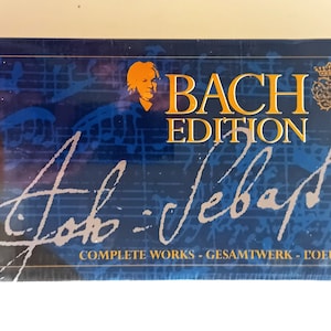 Op de afbeelding: Een blauwe doos met een portret van Johann Sebastian Bach en de tekst "BACH EDITION" en "Johann Sebastian Bach" in cursief. De doos bevat 155 cd's van Bach's complete werken.