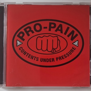 Op de afbeelding: Een vierkant CD-doosje met een felrode hoes. Het ontwerp bevat een zwarte ovale rand met de tekst PRO-PAIN bovenaan en CONTENTS UNDER PRESSURE onderaan, met een centrale afbeelding van een gebalde vuist en kleine driehoekjes.
