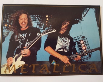 May include: A vintage trading card featuring a black and white photo of two musicians from the band Metallica playing electric guitars. The card has a gold border and the band's name "METALLICA" is written in gold lettering across the bottom.