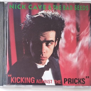 Op de afbeelding: Een compact disc hoesje met het album Kicking Against The Pricks van Nick Cave & The Bad Seeds. De hoes toont een portret van een persoon in een zwart pak met vlinderdas tegen een rode achtergrond met verticale zwarte strepen.