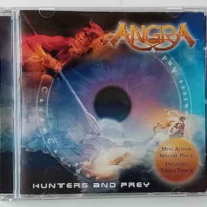 Puede incluir: Una caja de CD con un fondo azul y morado que presenta una figura roja sosteniendo una lanza. El texto "ANGRA" está en blanco y el texto "Hunters and Prey" está en negro. El texto "Mini Album Special Price Includes Video Track" está en blanco sobre un fondo rojo.