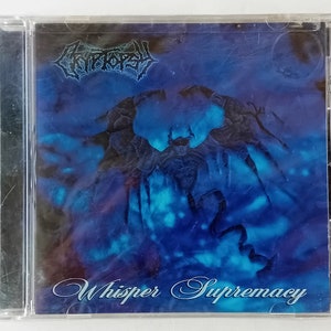 May include: A CD case with a blue and black cover featuring a dark figure with wings against a cloudy background. The text "Whisper Supremacy" is written in a cursive font at the bottom of the cover.