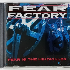 Puede incluir: Una caja de CD con un fondo negro y una imagen azul y blanca de engranajes. El texto "FEAR FACTORY" está en rojo y blanco, y el texto "FEAR IS THE MINDKILLER" está en rojo.
