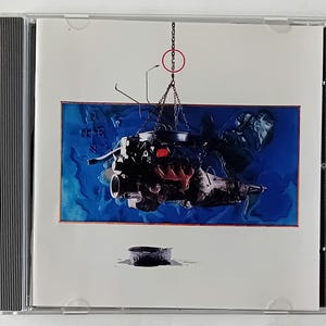 Può includere: Custodia di un CD con l'immagine di un motore di auto sospeso a una catena su sfondo blu. Il motore è nero con dettagli rossi. Un piccolo oggetto circolare scuro è posizionato sotto.