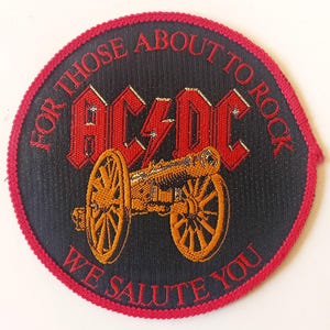 May include: A black and red embroidered patch with the text "For Those About To Rock We Salute You" and the band name "AC/DC" in red. A cannon with gold wheels is in the centre of the patch.