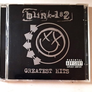May include: A Blink-182 Greatest Hits music CD in a clear plastic jewel case. The cover features a black background with a white smiley face logo, five arrows pointing in a circle, and a Parental Advisory Explicit Content label in the corner.