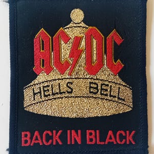 Könnte beinhalten: Ein schwarzer Stoff-Patch mit einer goldenen, gestickten Glocke mit der Aufschrift "Hells Bell". Die Band AC/DC ist in roten Buchstaben über der Glocke geschrieben. Die Worte "Back in Black" sind in roten Buchstaben unter der Glocke geschrieben.