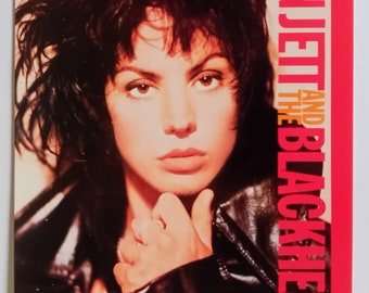 May include: A black and white photo of Joan Jett and the Blackhearts. Joan Jett is wearing a black leather jacket and has her hair styled in a short, choppy cut. She is looking directly at the camera with a serious expression.