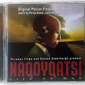 May include: A CD case with a red and white graphic of a robot-like figure with a sunset background. The text "NAQOYQATSI LIFE AS WAR" is printed on the front of the case. The text "Original Motion Picture Soundtrack music by Philip Glass cello solos by Yo-Yo Ma" is printed above the graphic. The text "Miramax Films and Steven Soderbergh present" is printed below the graphic.