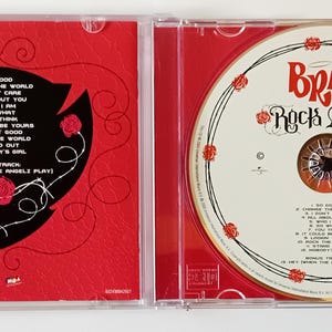 Puede incluir: Un CD de m&uacute;sica Bratz Rock Angelz en una caja de pl&aacute;stico transparente. El lado izquierdo presenta un fondo rojo con un escudo negro que contiene una lista de canciones. El lado derecho muestra el disco CD blanco con el logo de Bratz Rock Angelz.