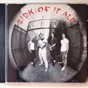 Op de afbeelding: Een muziek-cd-hoesje met een zwart-wit fisheye-foto van vier personen die in een steegje met gaashekken staan. De tekst SICK OF IT ALL staat in rode, grillige handgeschreven letters bovenaan de hoes gedrukt.