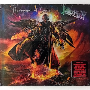 Puede incluir: Una portada de CD para el álbum "Redeemer of Souls" de Judas Priest. La portada presenta una figura oscura con alas, de pie en un paisaje de fuego. El título del álbum está escrito en una fuente gótica en negrita.