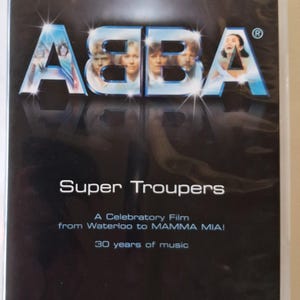 Pode incluir: Capa de DVD de "Super Troupers", um filme sobre ABBA. A capa apresenta o logotipo da banda em azul e prata, com imagens dos membros. O texto inclui "30 anos de música."