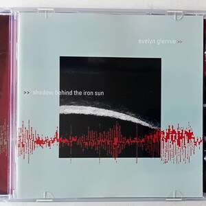 Può includere: Una custodia per CD con sfondo blu chiaro e un'immagine in bianco e nero di un orizzonte. Il testo "shadow behind the iron sun" è stampato in nero sulla parte anteriore della custodia. Il nome dell'artista, "evelyn glennie", è stampato in nero nell'angolo in alto a destra della custodia.