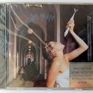 Puede incluir: Una caja de CD para el álbum "Pink Bubbles Go Ape" de la banda "Helloween". La portada muestra a una mujer con un vestido blanco sosteniendo un pez en un pasillo con arcos.