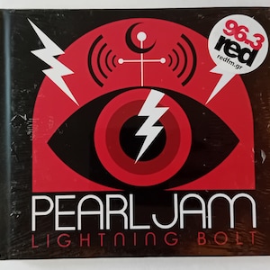 Puede incluir: Una caja de CD roja con un gráfico en blanco y negro de un ojo con un rayo dentro. El texto "PEARL JAM LIGHTNING BOLT" está impreso en blanco en la parte delantera de la caja. Una pegatina roja con el texto "96.3 red redfm.gr" está en la esquina superior derecha.