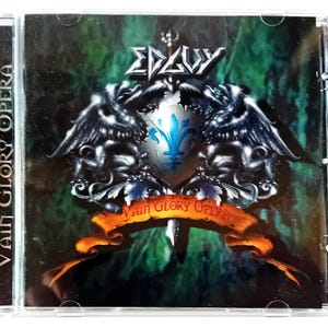 Op de afbeelding: Een cd-hoesje met het album "Vain Glory Opera" van Edguy. De hoes toont een schild met een fleur-de-lis, gevleugelde figuren en een banner met de albumtitel. De achtergrond is een mix van groen en donkere kleuren.