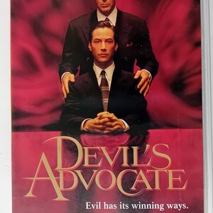 May include: A VHS tape cover for the movie "Devil's Advocate" starring Keanu Reeves and Al Pacino. The cover features a red background with the title in gold lettering. The tagline is "Evil has its winning ways." and the movie is rated 18.