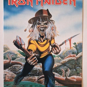 Può includere: Un poster rettangolare con il testo IRON MAIDEN in alto. L'illustrazione raffigura una figura scheletrica con un cappello marrone e una maglietta gialla, che impugna un'ascia e un coltello insanguinati mentre è immersa in acqua tra rocce.