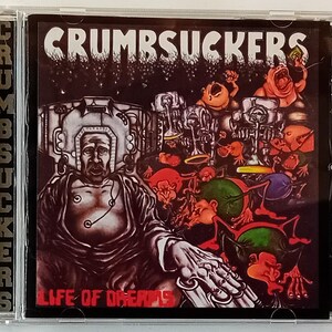 Op de afbeelding: Een CD-hoes met een kleurrijke, psychedelische albumhoes met een cartoonachtige figuur met een groot hoofd en een donkere achtergrond. De albumtitel is "Crumbsuckers - Life of Dreams".