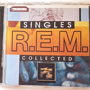 Op de afbeelding: Een doorzichtig plastic CD-doosje met het album Singles Collected van R.E.M. De hoes bevat een collage in blauwe en oranje tinten, met de tekst SINGLES en R.E.M. COLLECTED in wit en zwart, plus een kleine code 243.8.29642.23.