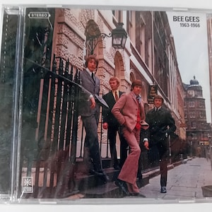 Puede incluir: Una caja de CD con una foto de los miembros de la banda Bee Gees de pie en una calle de la ciudad. Los miembros de la banda visten ropa de la década de 1960. La caja de CD tiene el texto "Bee Gees 1963-1966" impreso en la parte delantera.