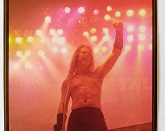May include: A man with long blonde hair is singing into a microphone on stage. He is wearing black trousers and a black sleeveless shirt. The stage is lit with red and yellow spotlights.