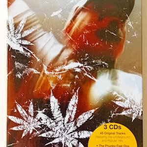 May include: A 3-CD collection of Cypress Hill music. The cover features a close-up of a man's face with a baseball cap on, and a marijuana leaf design in the background. The text on the cover reads "Cypress Hill the collection" and "Presenting 3 Original Album Classics".